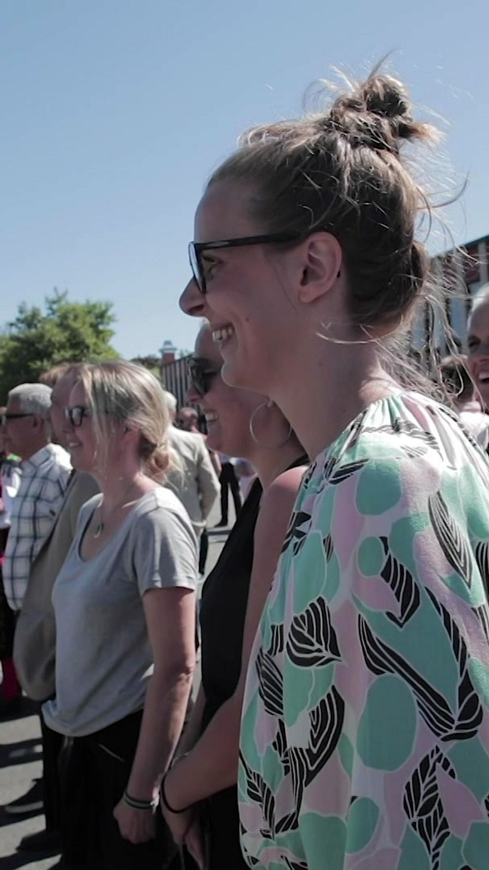 En grupp människor står samlade utomhus vid en skolgård med en svensk flagga hissad i bakgrunden, det är en solig dag.