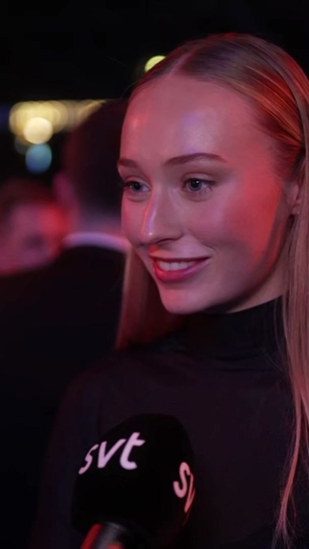 En kvinna i en svart outfit står i fokus för en intervju på en gala. Bakgrunden visar suddiga silhuetter av människor och färgglada ljuspunkter som skapar en festlig atmosfär.
