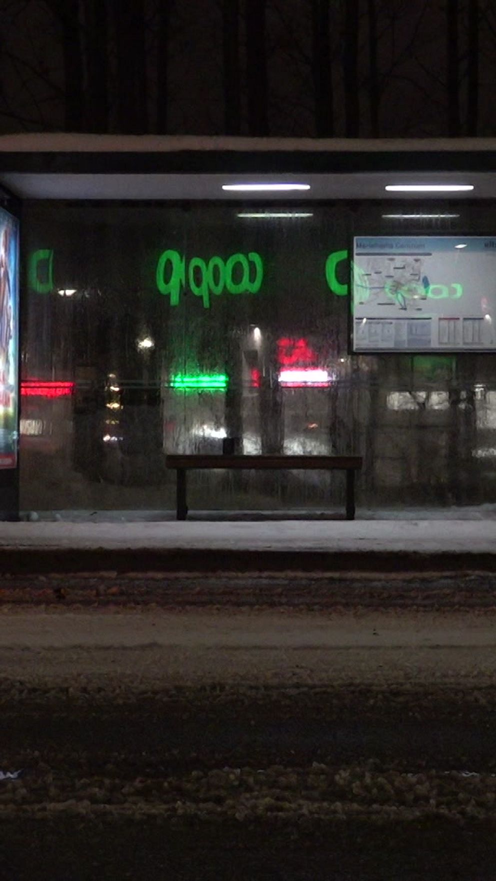 En busshållplats i mörker med snö på marken, där en bänk står under ett tak. Bakgrunden visar en ICA-butik med lysande skyltar och reklamer.
