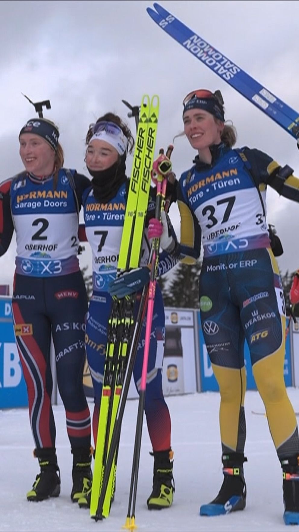 Tre skidåkare poserar tillsammans efter en tävling, håller sina skidor i luften. De bär tävlingsdräkter och står på snöig mark, med sponsormärken och en blå DKB-bakgrund i synfältet. Runt omkring dem syns fler åkare och en vinterlandskap.