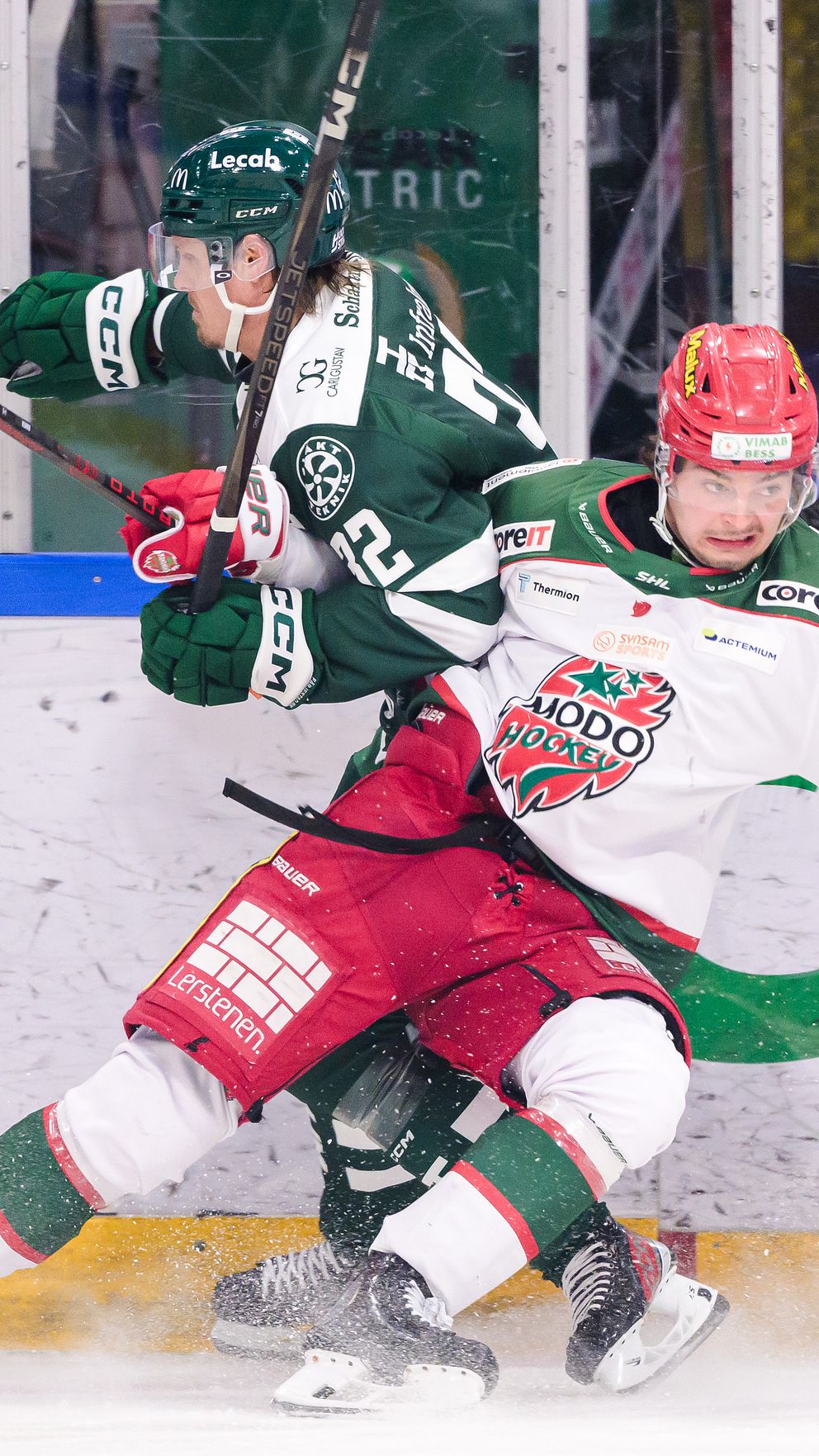 Två ishockeyspelare i intensiv kamp på isen, där en spelare från laget i gröna tröjor blir tacklad av en motståndare i vit och röd tröja. I bakgrunden syns åskådare och delar av ishallarens inredning.
