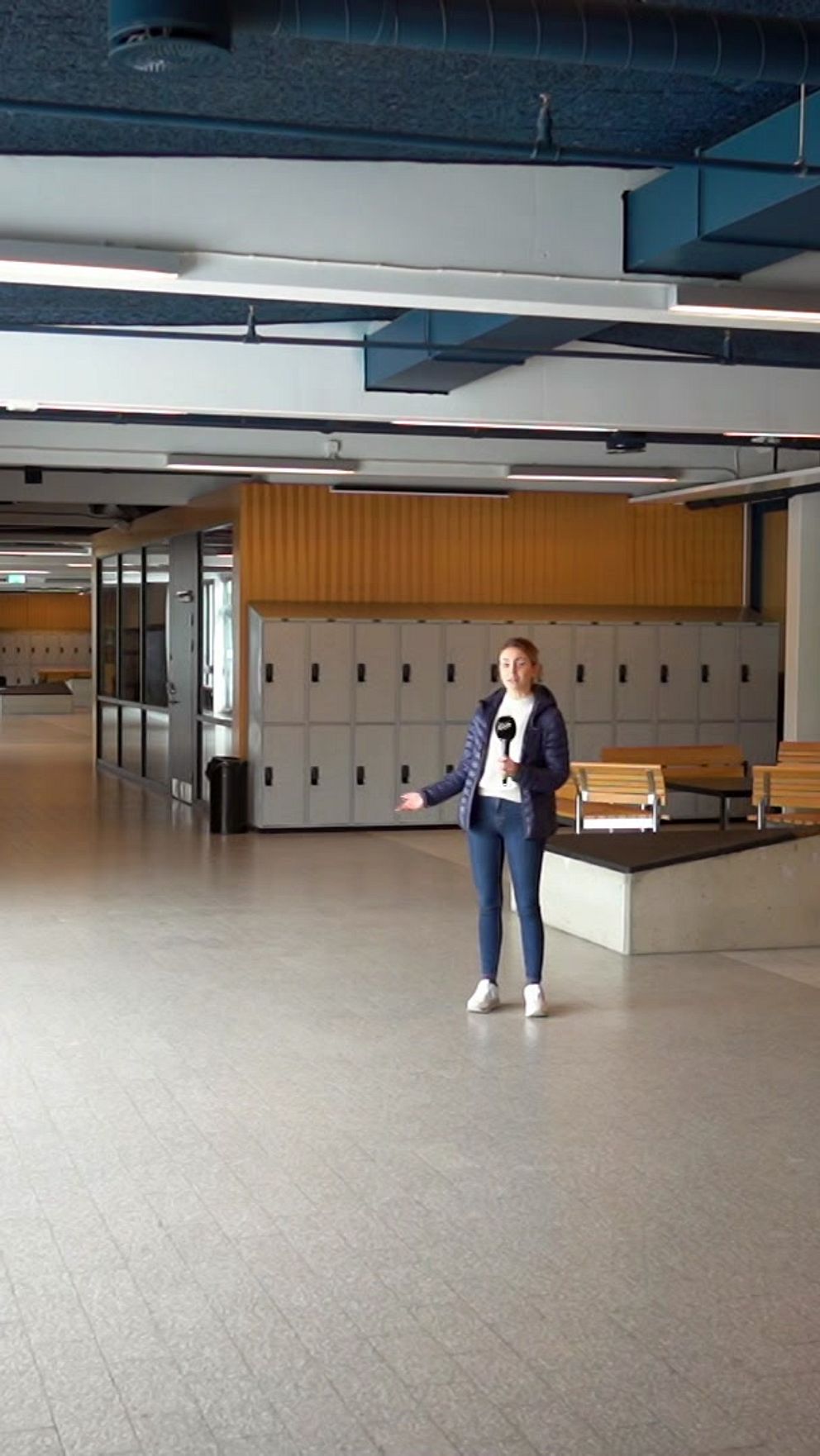 En kvinna står mitt i en lång, bred korridor med ljusa golv och högt i tak. Till höger finns en rad skåp och en dörr märkt ”WC”. Korridoren är tom förutom personen, och det finns bänkar längs fönstren till vänster.
