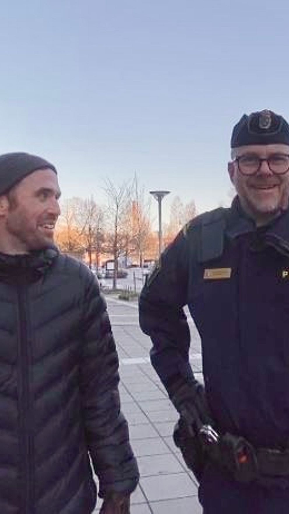 En person i en mörk jacka står bredvid en polisofficer i uniform utomhus. Scenen utspelar sig på en plats med moderna byggnader i bakgrunden och snö på marken. Det är en klar dag med lite ljus som reflekteras i fönstren på byggnaderna.