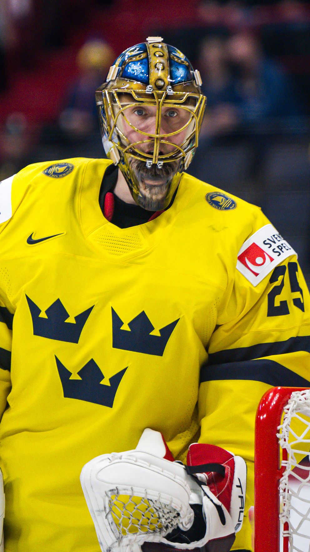 Målvakt i Tre Kronor-tröja står vid ishockeymålet under match med publik i bakgrunden.