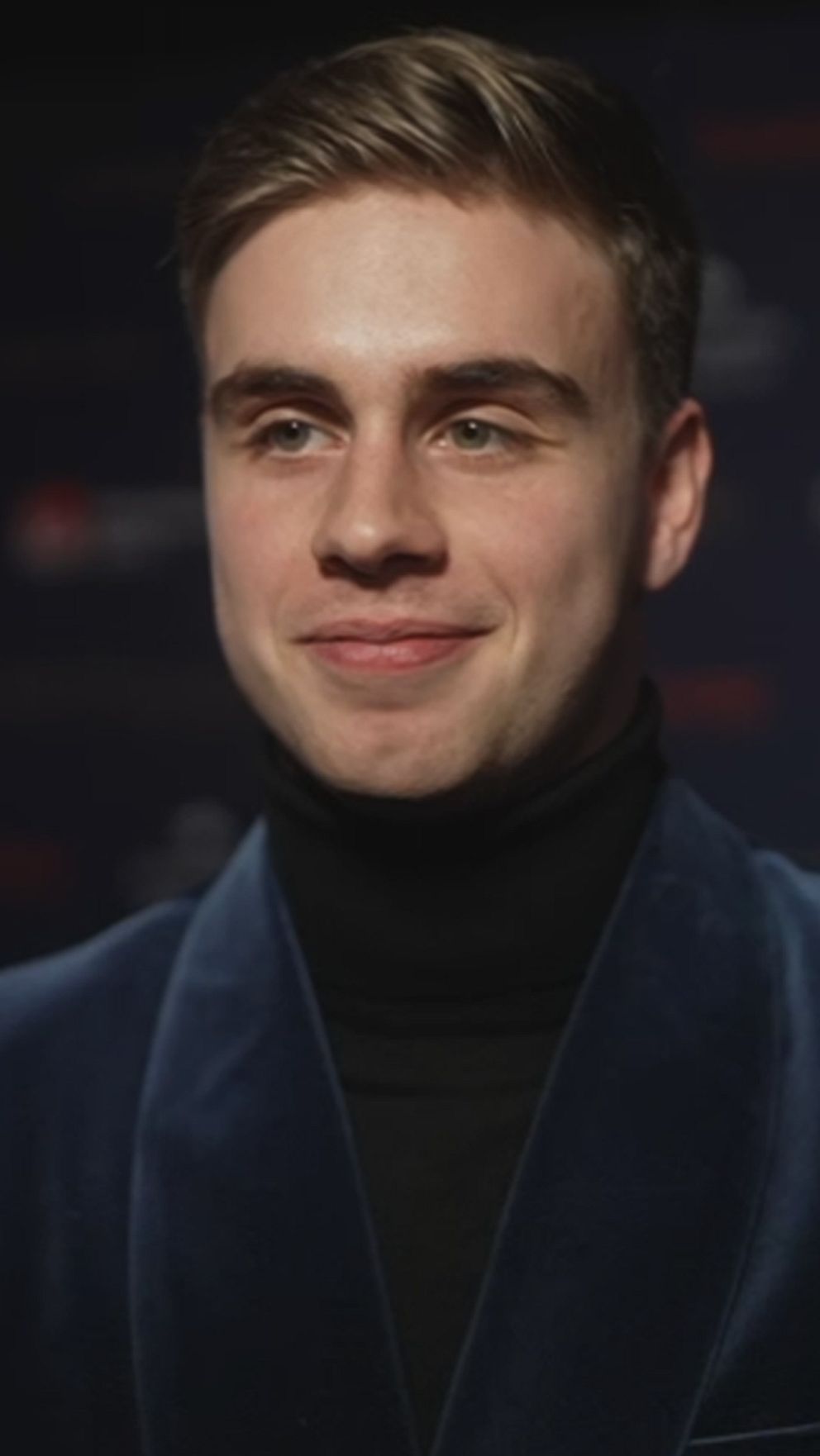 En person iklädd en blå sammet blazer står framför en mörk bakgrund med diskreta logotyper, troligtvis på en gala eller ett offentligt evenemang.