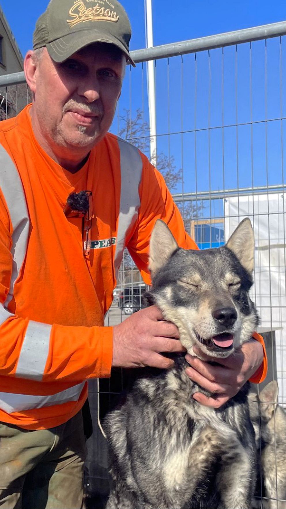 Person i orange varselkläder klappar en hund vid byggarbetsplats.