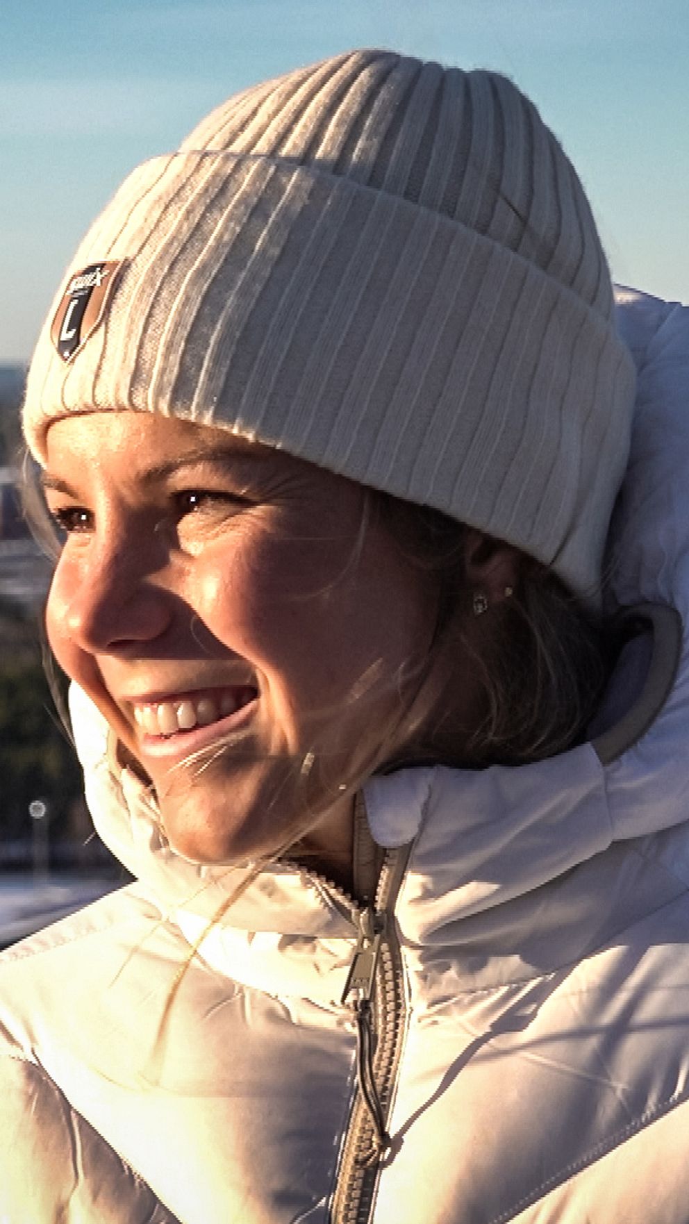 Längdskidåkaren Johanna Hagström iklädd en vit jacka och en ruta med en stickad mössa står vid en utsiktsplats i Falun. Bakgrunden visar en vinterlandskap med snö, träd och en fjärran vy av täckta kullar och en sjö. Dagsljuset skapar en klar och kall atmosfär.