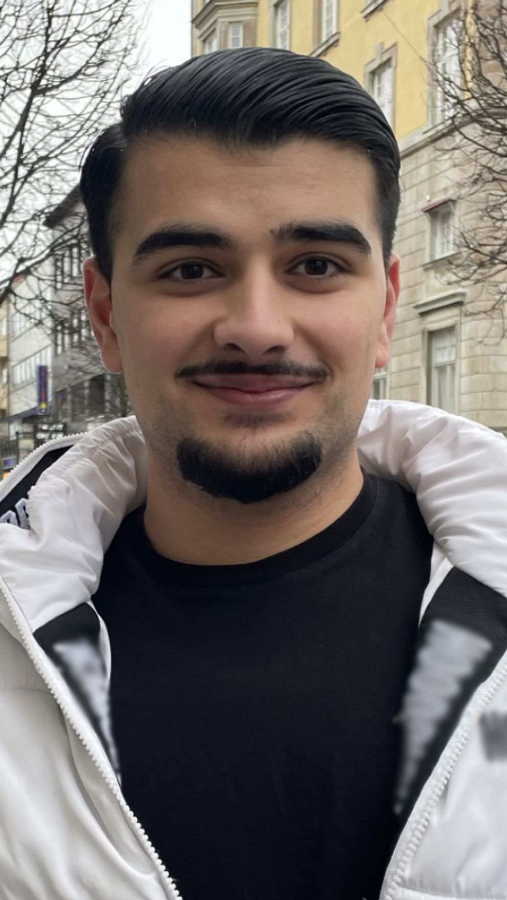 En man iförd en vit jacka står på en trottoar. Bakom syns en lång gata med butiker och restauranger.