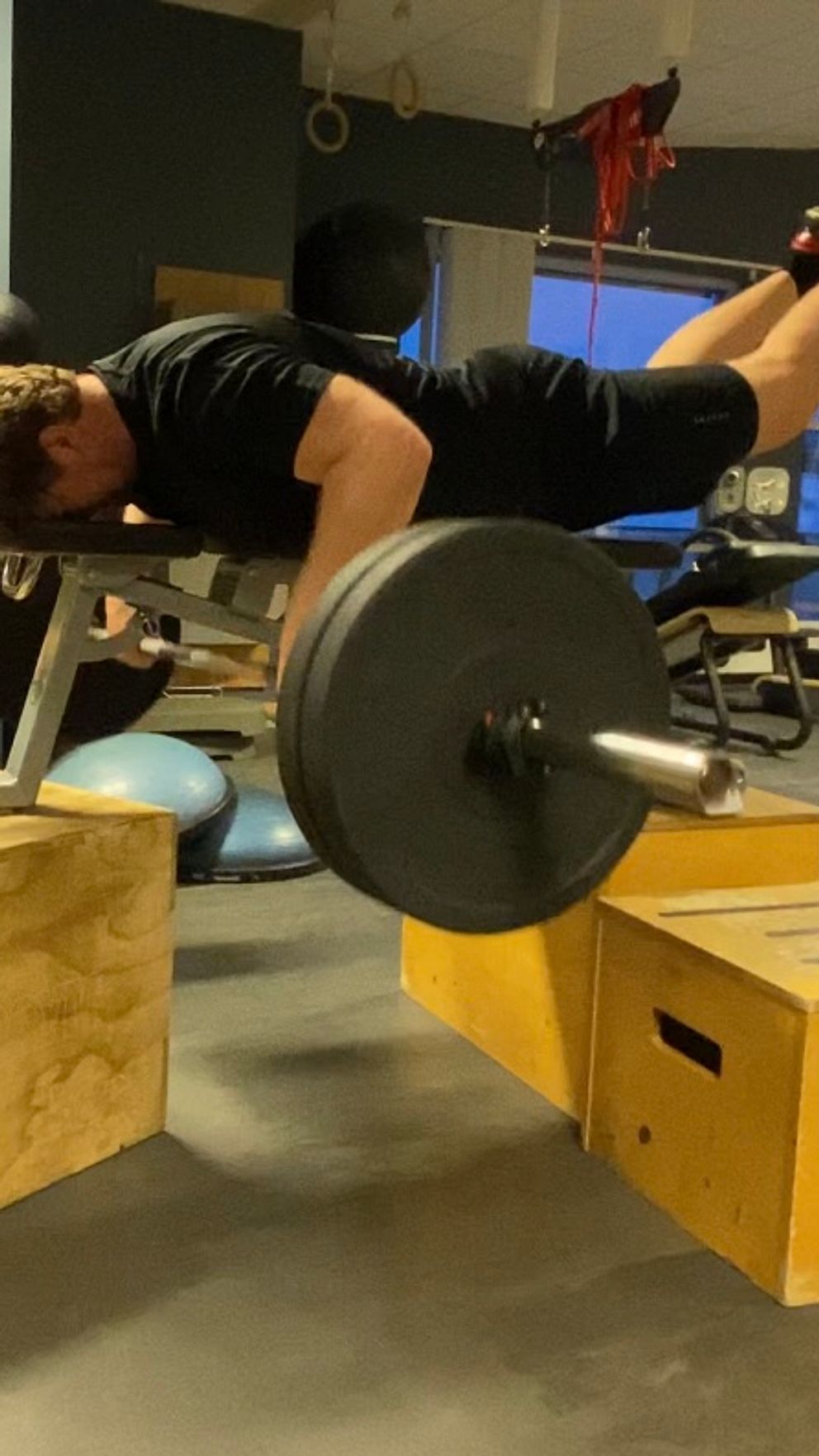 En person lyfter en tung skivstång medan hen balanserar magläge på en bänk som stöds av två trälådor i ett gym. I bakgrunden syns träningsutrustning och ett fönster.