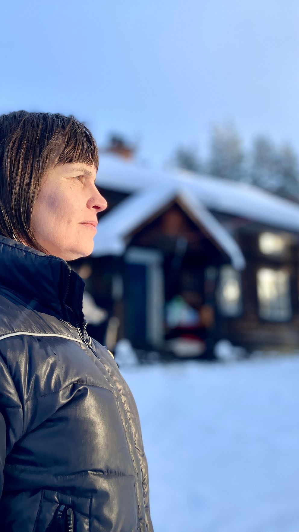 En person i vinterkläder står utomhus med ett snötäckt trästuga i bakgrunden, omgiven av snöpudrade träd. Johanna Karlsson bor offgrid utanför Delsbo.