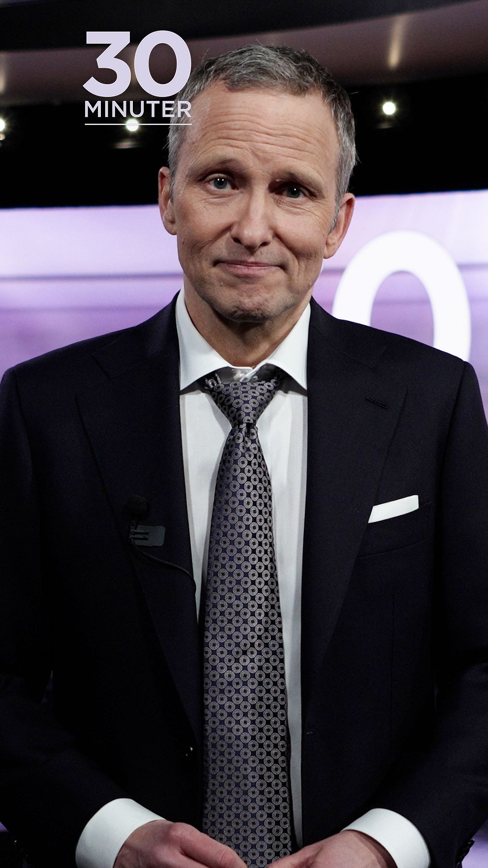 Anders Holmberg i kostym med 30 minuter-logotyp, TV-studiobakgrund.