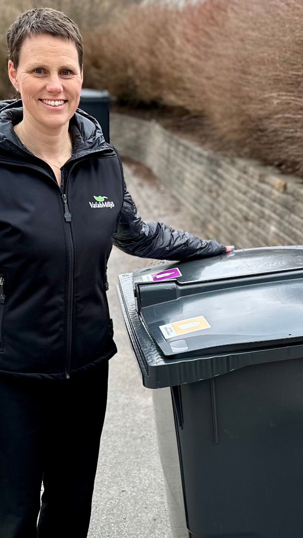 Person står bredvid en sopcontainer på trottoar. Lägre buskar längs en stenmur.