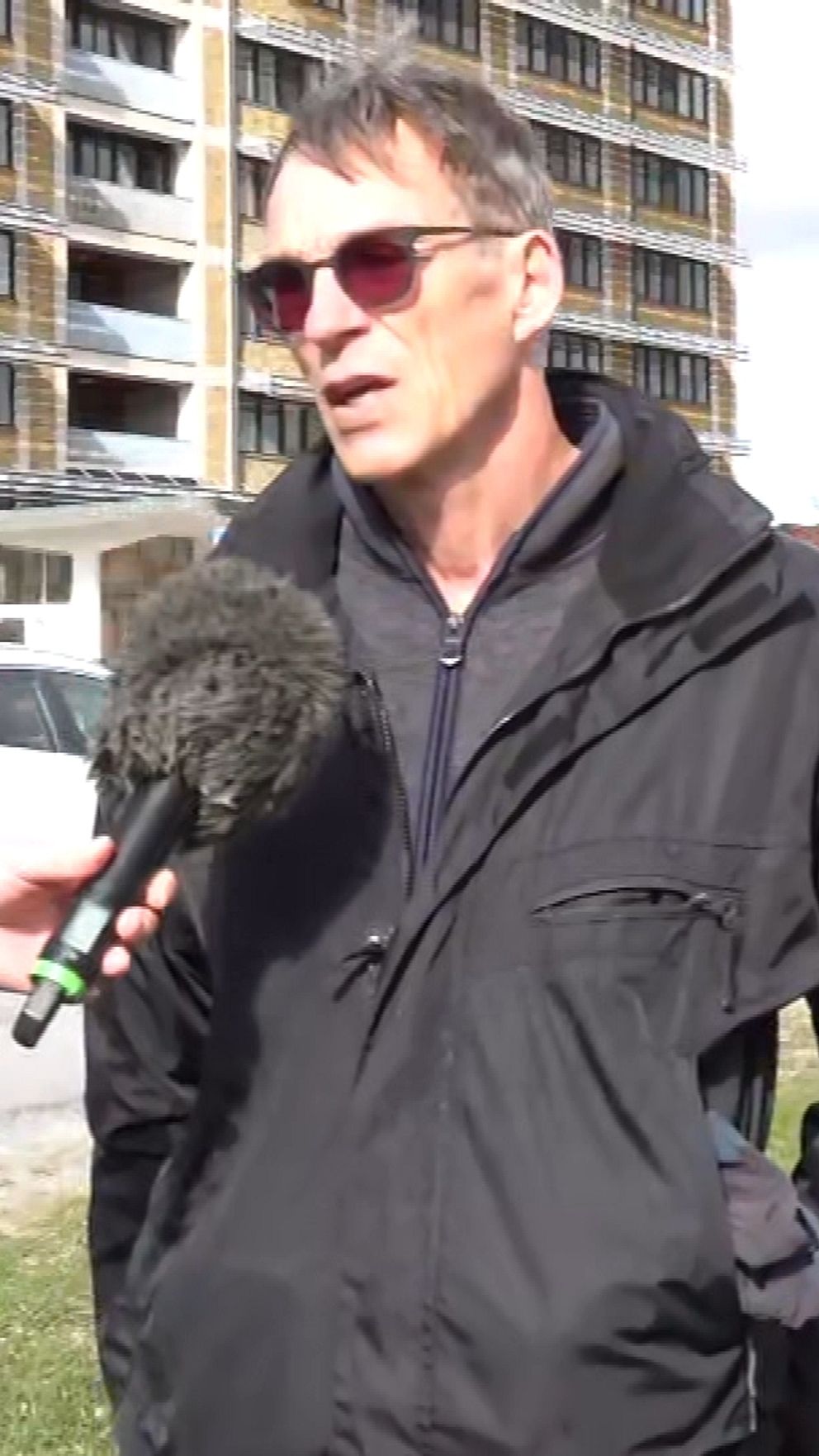En reporter intervjuar en person utomhus framför ett bostadshus och parkerade bilar.