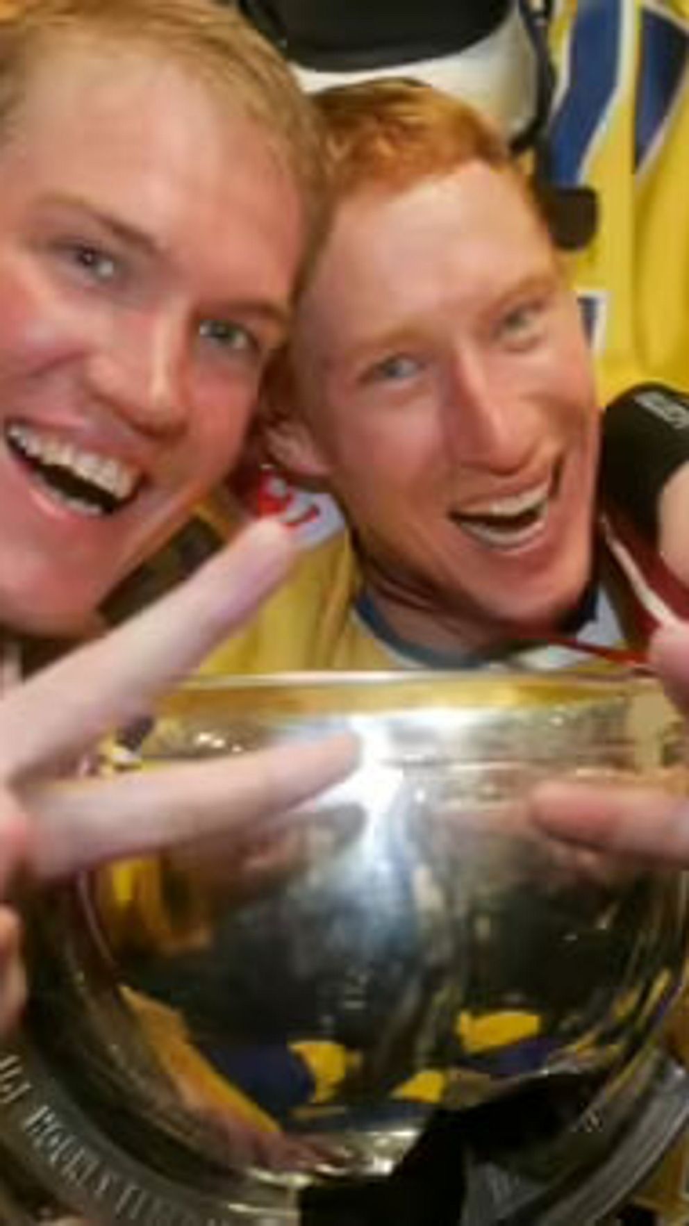 Två personer i svenska hockeytröjor firar seger med pokal och segertecken i omklädningsrum.