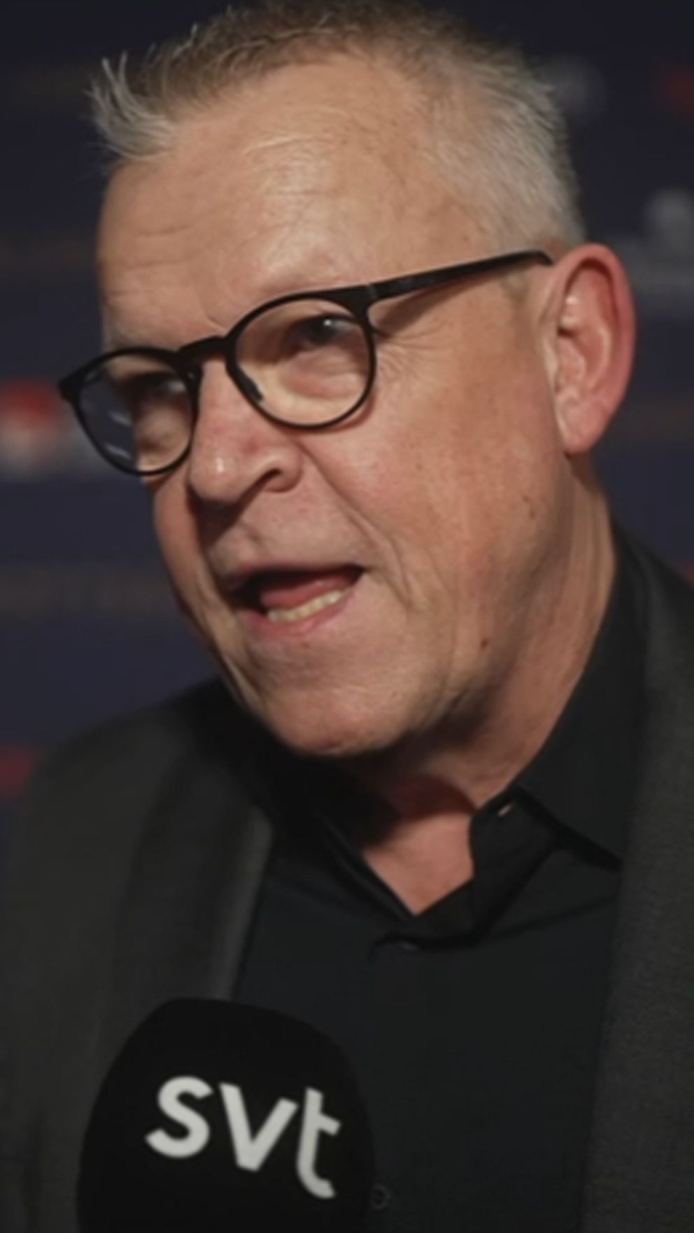 En man i svart kostym och mikrofon intervjuas på en rödmatta-event, omgiven av personer i bakgrunden.
