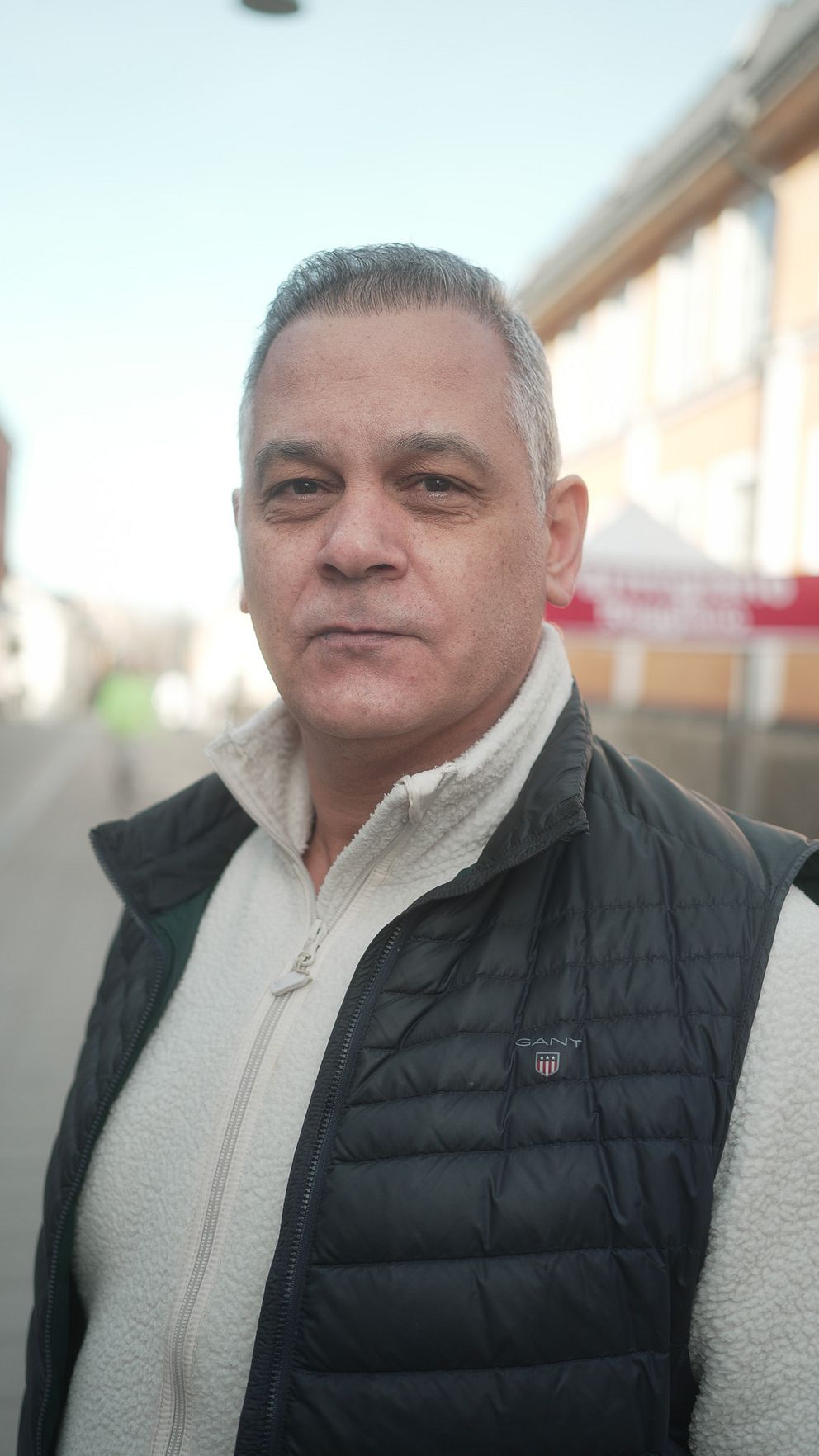 Person står på en trottoar, iklädd vit tröja och svart väst. Stadsmiljö i bakgrunden.