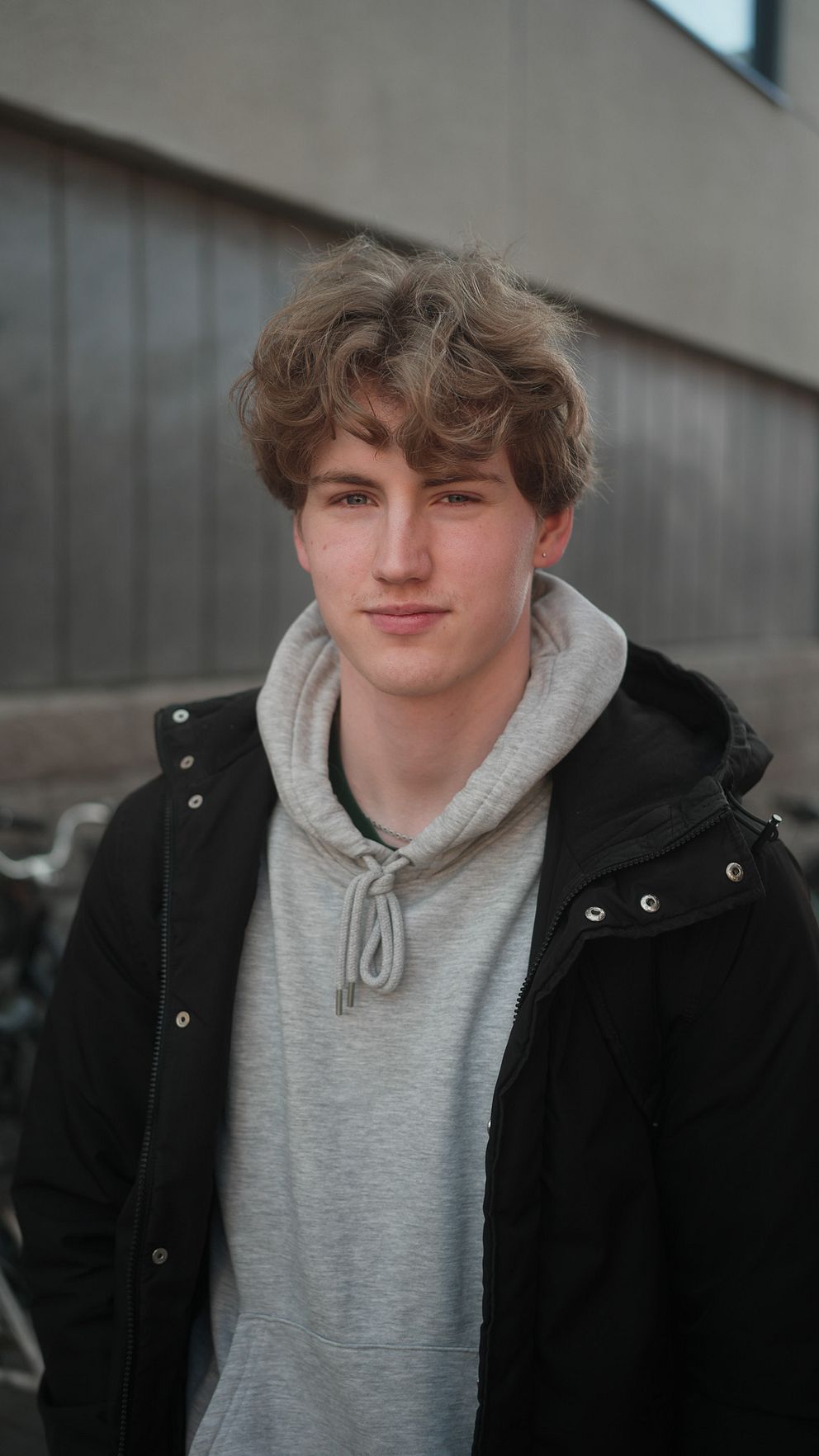 Person i grå hoodie och mörk jacka framför grå byggnad.