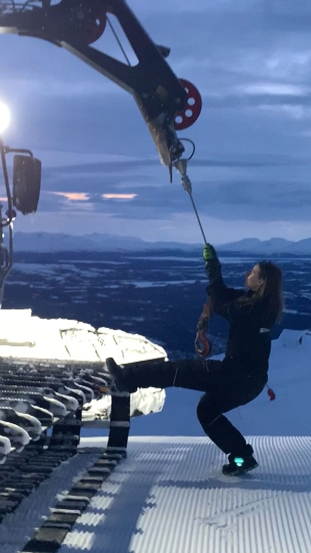 Pistmaskin i fjällmiljö med en person som håller fast i en vinsch, mot en skymningshimmel.