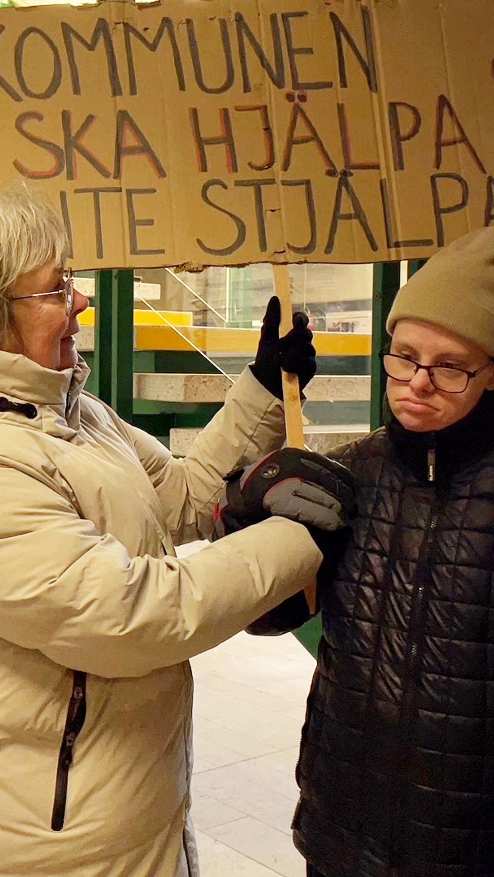 Två personer håller upp en skylt med texten: ”Kommunen ska hjälpa, inte stjälpa”