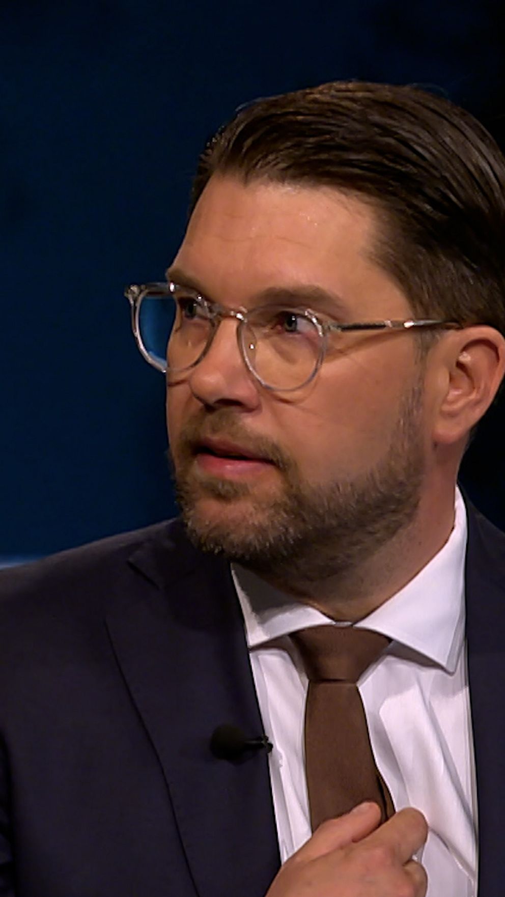 Man i kostym och slips deltar i en tv-intervju med mörk bakgrund och ljusa ränder.
