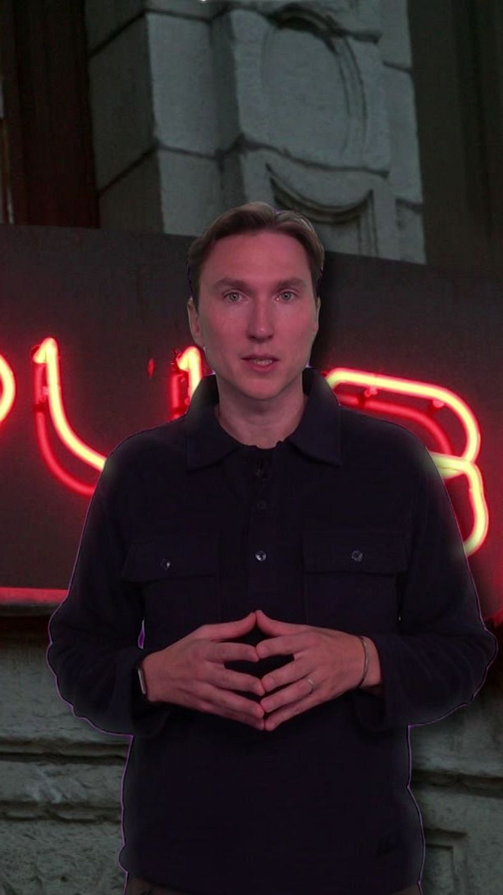 En person står framför en ljus, röd neonskylt med texten ”THE SPY BAR”. Bakgrunden visar en arkitektonisk fasad, vilket ger en urban atmosfär.