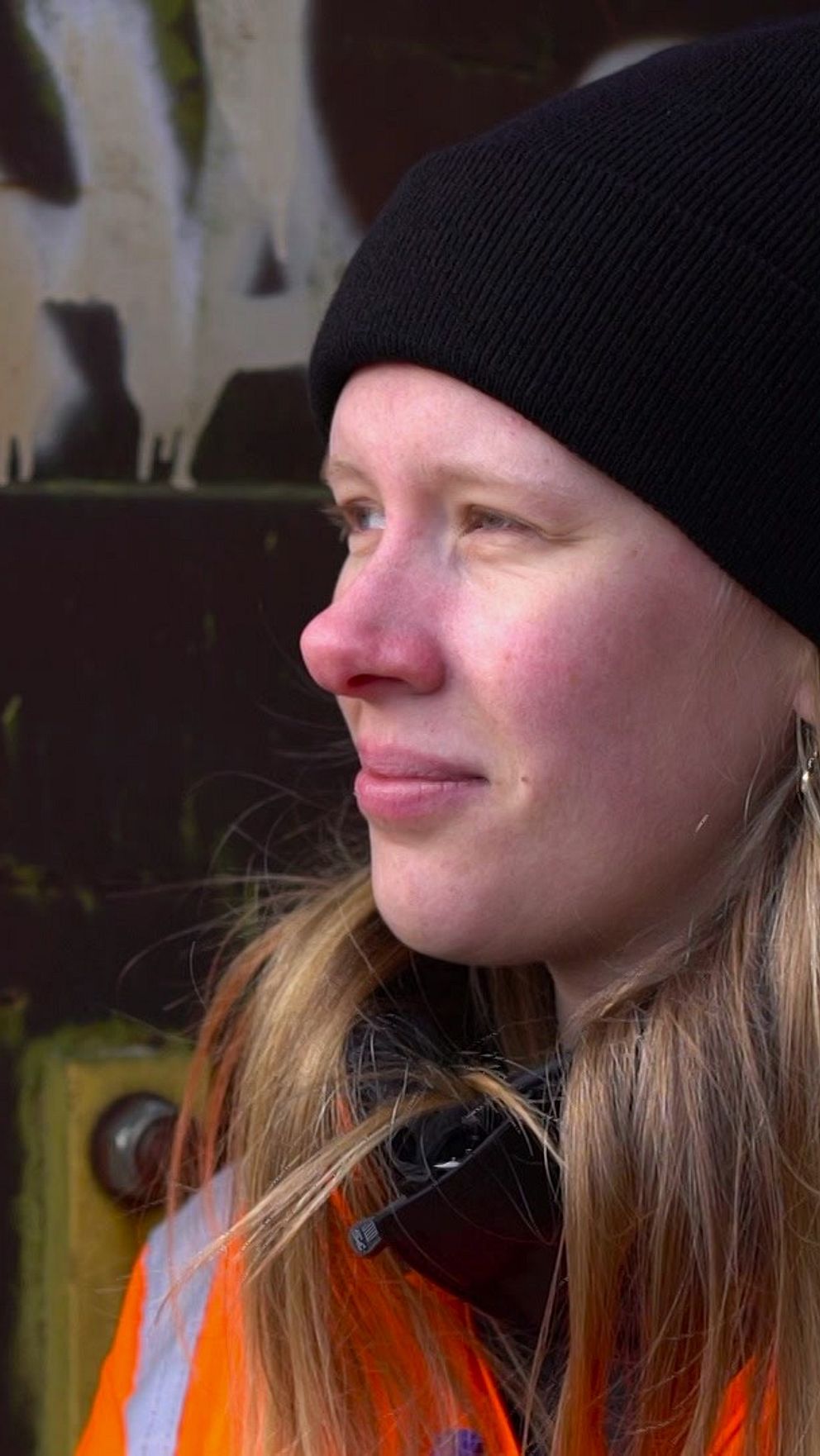 Person i orange arbetsjacka, framför en graffiti-prydd yta.