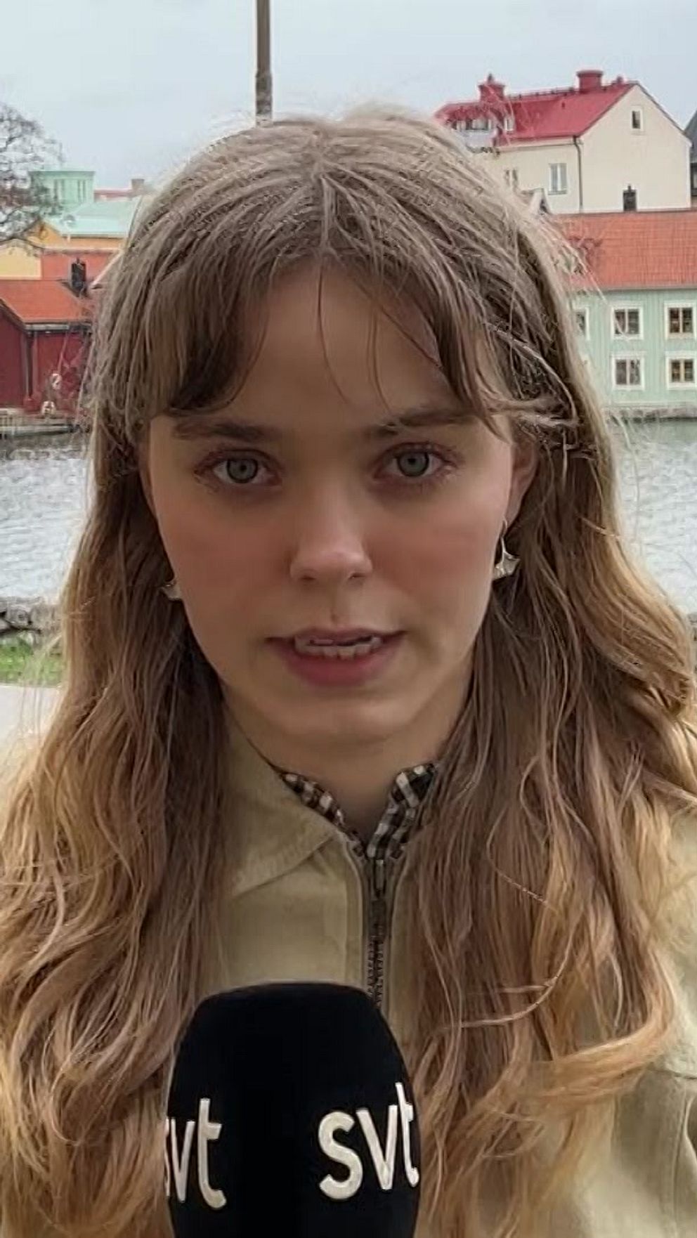 Reporter med SVT-mikrofon framför en å vid färgglada hus, en person promenerar i bakgrunden.