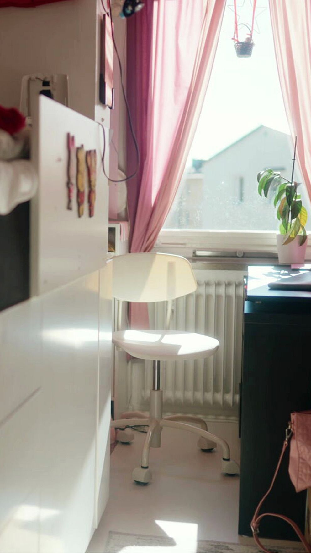 Rummet har en säng på en övre nivå, med en vit stol i fokus i ljusstrålar från fönstret. Mörka möbler, inklusive en låda, syns bredvid stolen, och det finns en rosa väska på golvet. Fönstret är täckt med rosa gardiner, och en växt står i fönstret.