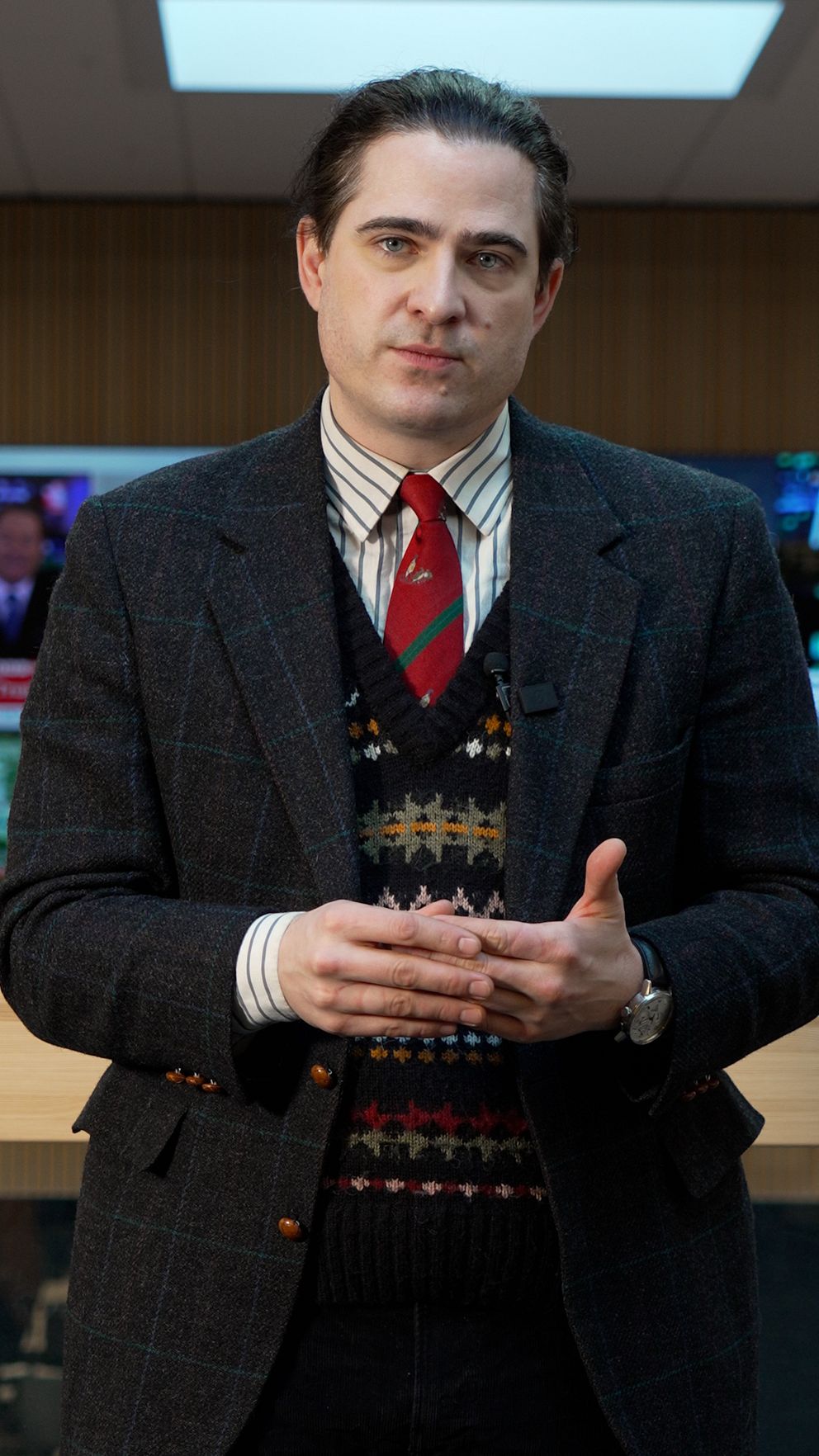 Man i kavaj och slips står framför TV-skärmar, händerna sammanflätade.