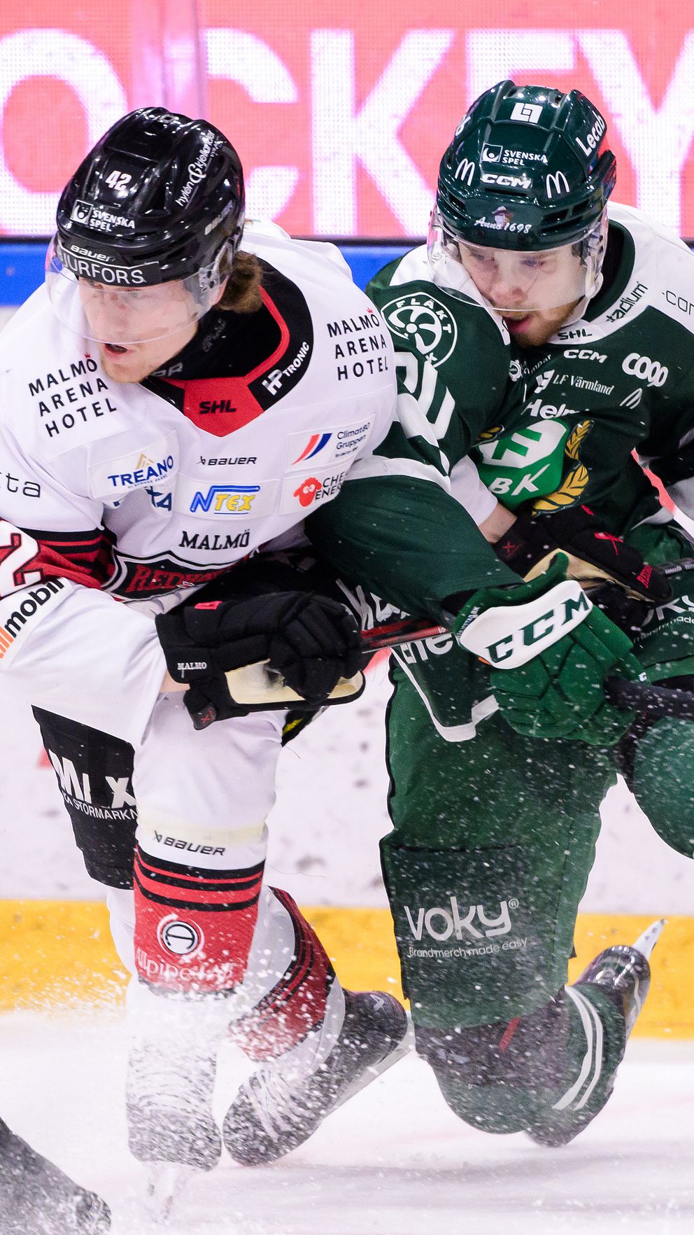 Två ishockeyspelare är engagerade i en närkamp på isen medan en tredje spelare observerar från vänster. Den ena spelaren bär en vit tröja med röd och svart detalj, medan den andra har en grön tröja. Bakgrunden visar en stor reklamskylt som annonserar för ishockey.