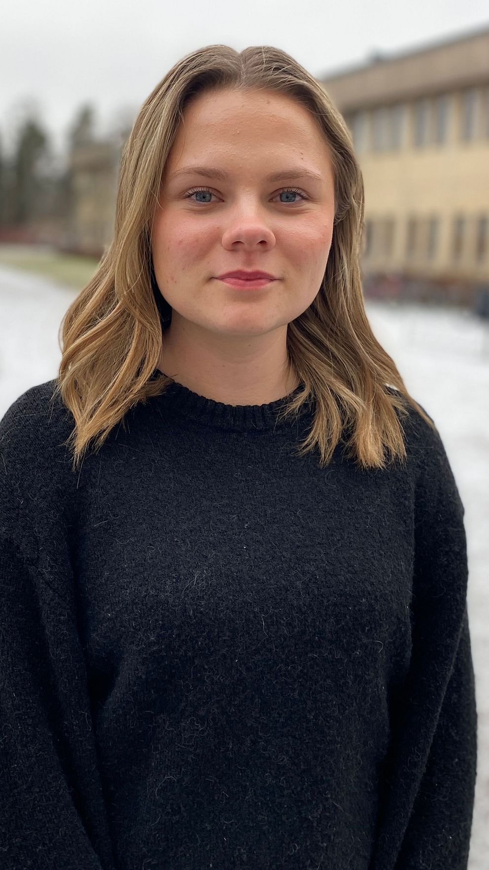 En person står utomhus på en snötäckt yta framför en lång byggnad, troligtvis en skola eller kontor, med träd i bakgrunden.