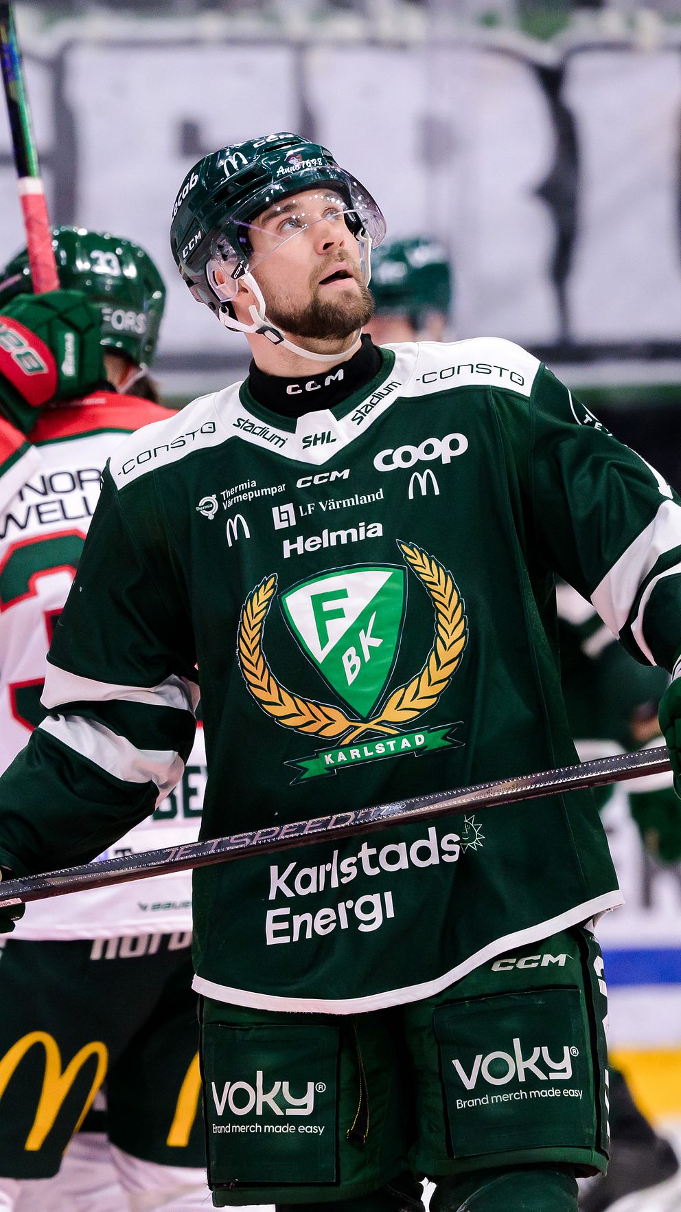 En hockeyspelare i grön tröja ställer sig med sin klubba i handen efter att ha gjort mål, medan lagkamrater i röda tröjor firar i bakgrunden.