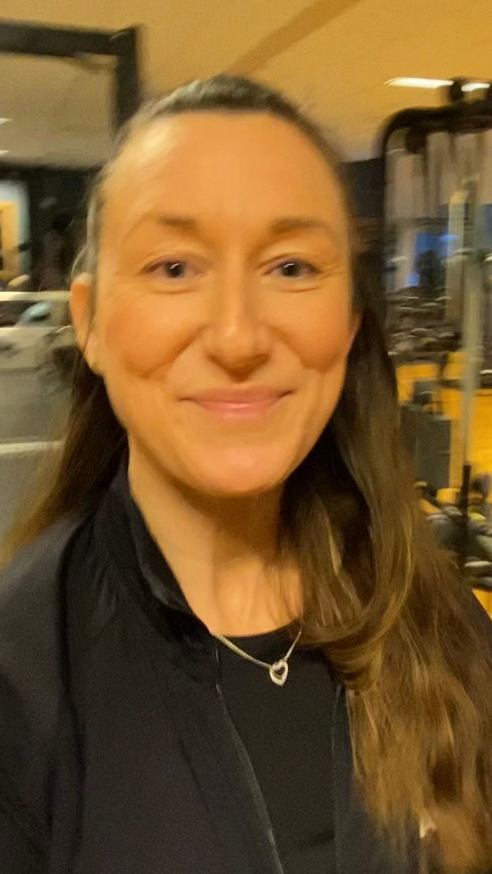 En kvinna står i ett gym fyllt med träningsutrustning som exempelvis vikter och maskiner, belyst med varma lampor.
