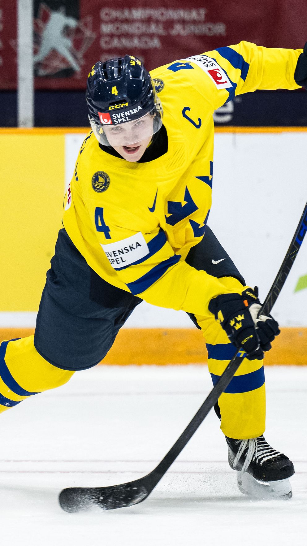Svensk ishockeyspelare i gult lagtröja gör ett skott på pucken under en junior-VM-match. Designsatsning i bakgrunden med sponsorlogotyper och arenainformation.