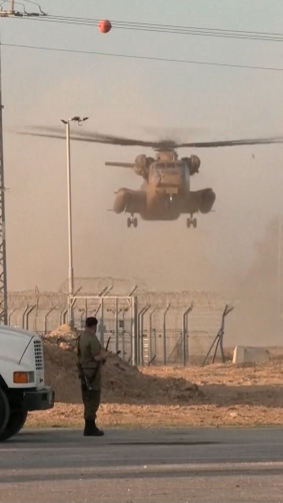 Ett militärhelikopter lyfter från en bas medan en soldat står vid två militärlastbilar i förgrunden, omgiven av damm och en staketstruktur med taggtråd i bakgrunden.