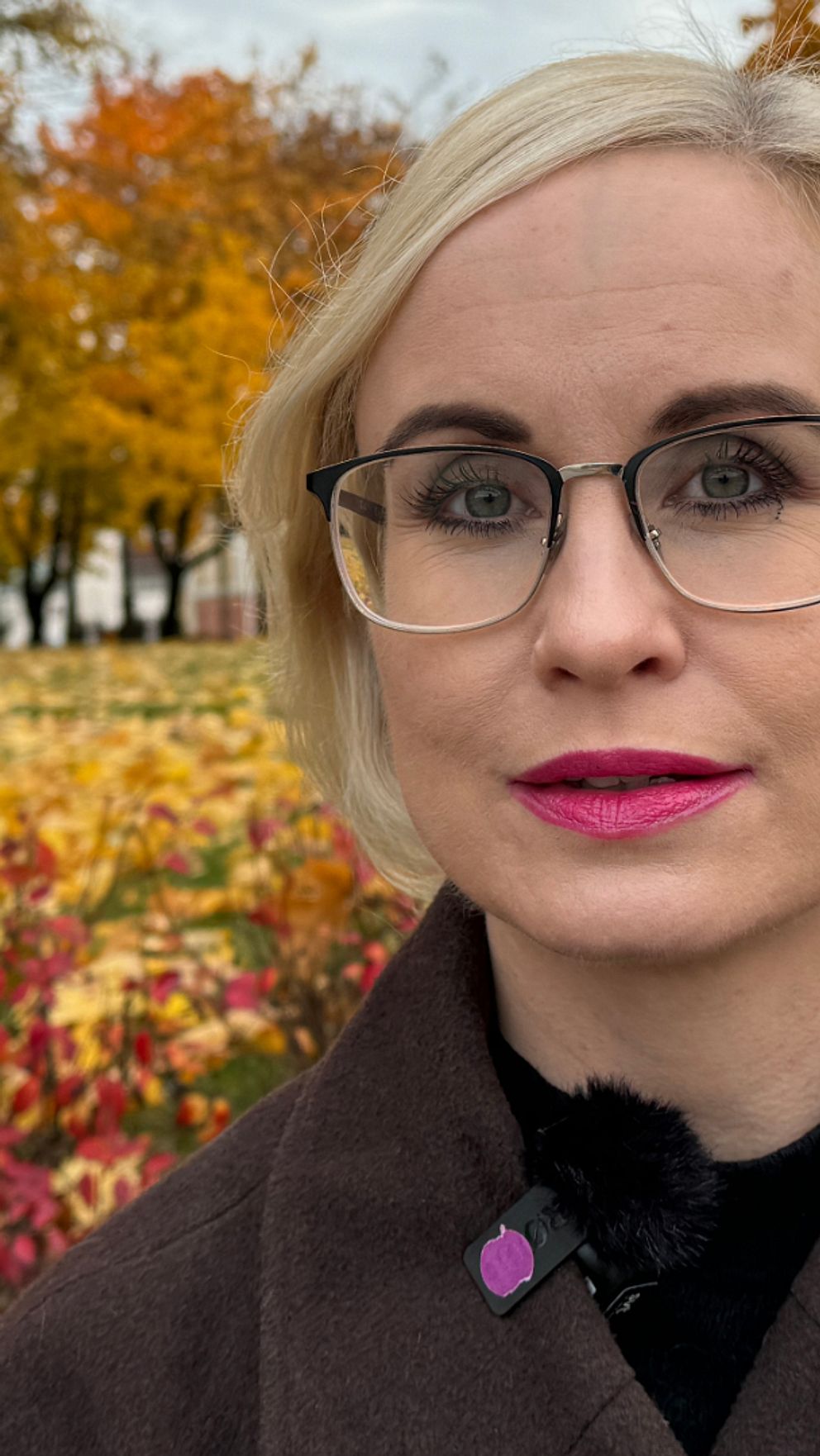 Kammaråklagare Karolina Lassbo står i en park med höstliga träd och färgglada löv på marken. Bakgrunden visar en tydlig höstscen med orange och gula löv samt en byggnad i avlägsen höger del av bilden.