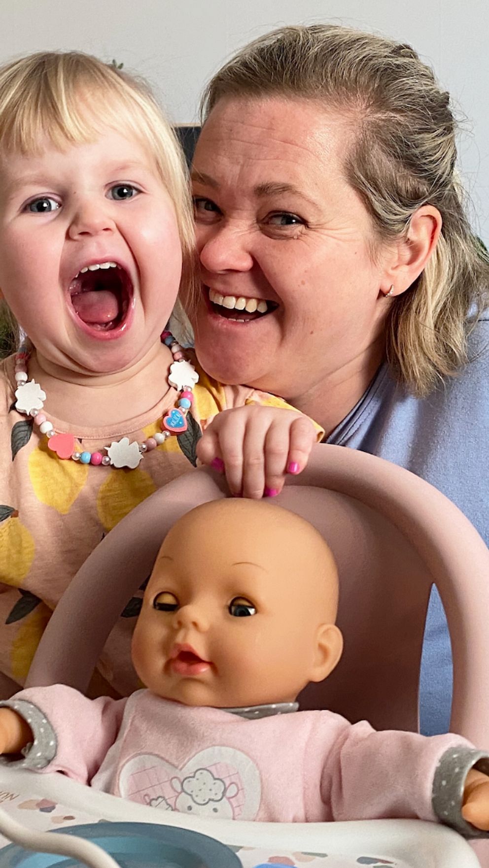 En liten treåring, thea, och hennes mamma erica, ler in i kameran, stort, och framför sitter en docka i en barnstol.