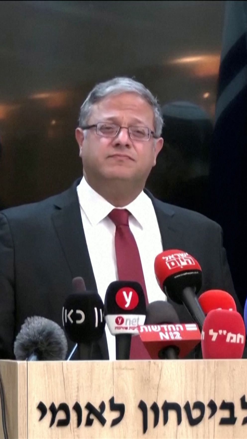 Tre personer med kippa står vid ett podium med mikrofoner och medieutrustning under en presskonferens. I bakgrunden syns en israelisk flagga och en skylt med hebreisk text, vilket indikerar att evenemanget äger rum i Israel. Personerna är formellt klädda i kostymer med en av dem i röd slips.