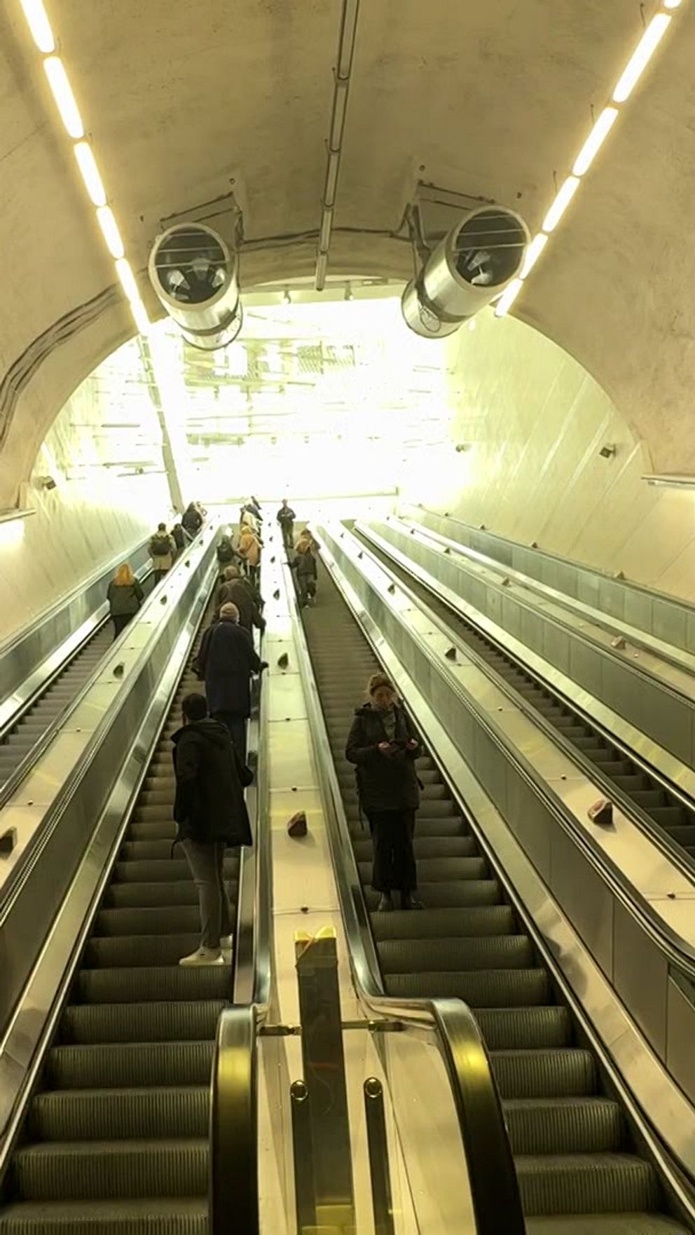 En stort, modernt rulltrappa i en tunnelbaneplattform som leder uppåt mot ett ljust område. Människor färdas upp och ner i rulltrappan, medan den omgivande strukturen har en stenig och industriell design med stark belysning.
