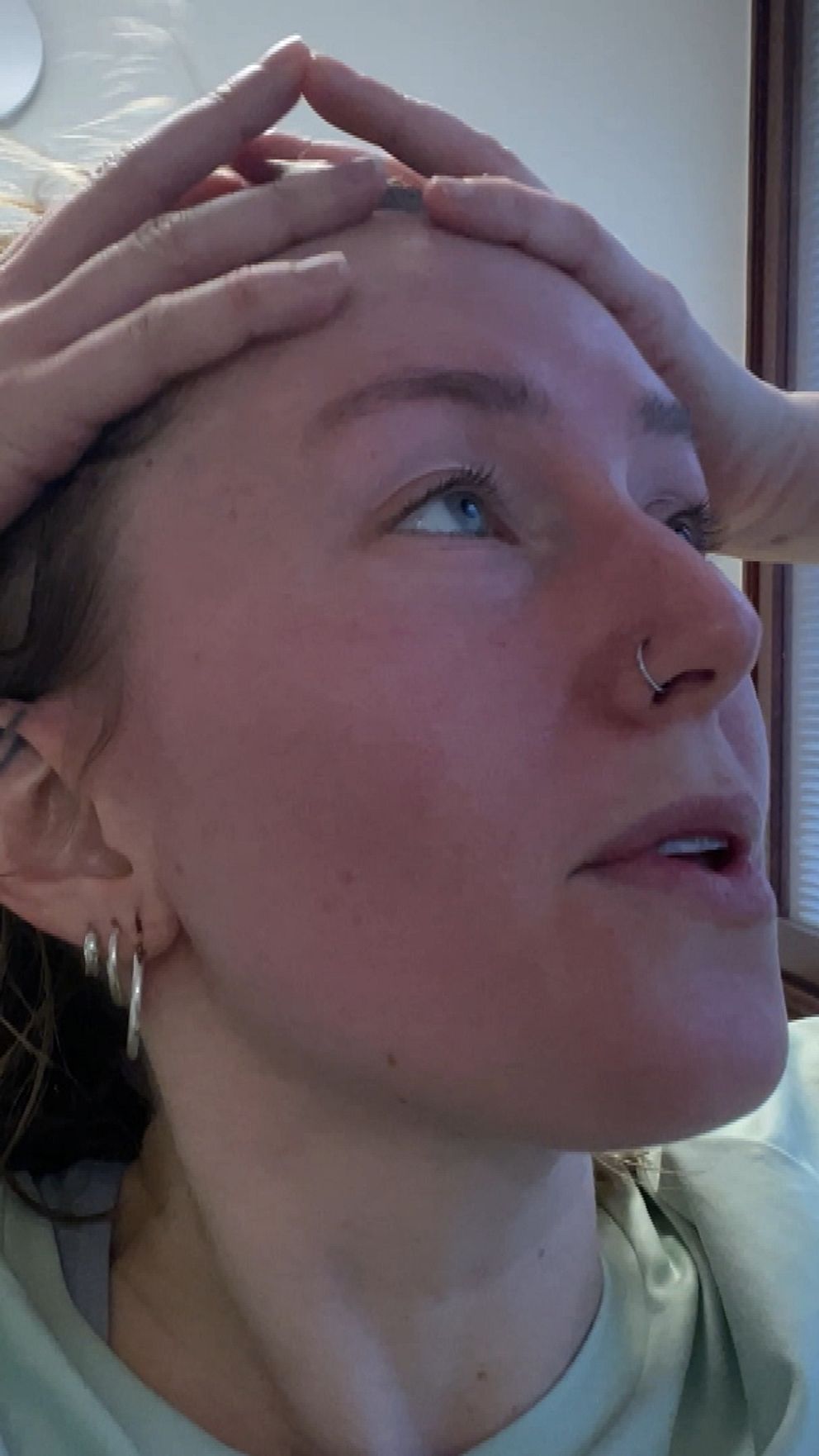 En person står i ett rum med en grön tröja och håller sina händer på huvudet. I bakgrunden syns en del av rummet med ett fönster och en dörr.
