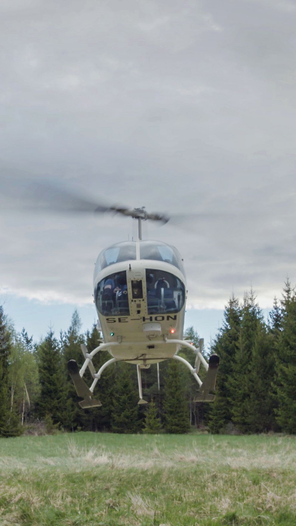En helikopter svävar över en öppen gräsmark, omgiven av tät skog. Himlen är delvis molnig, vilket skapar en dramatisk bakgrund till scenen.
