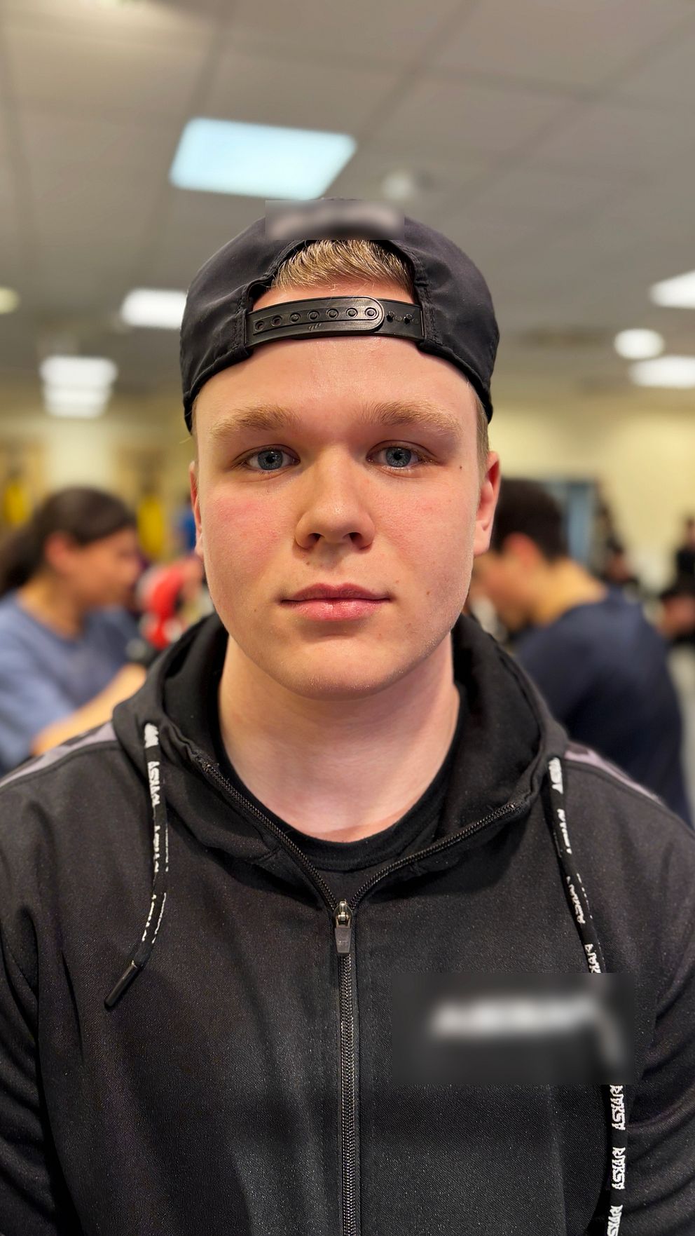 Person i svart hoodie står i en gymhall på Västerås fight club på Bäckby; flera människor i bakgrunden.