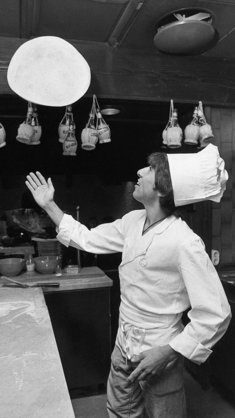 En kock står i en restaurangkök och kastar en rund degplatta i luften. I bakgrunden hänger olika köksredskap och ingredienser, medan flera skålar står på bänken framför kocken. Kockens klädsel inkluderar en vit kockrock och en traditionell kockmössa.