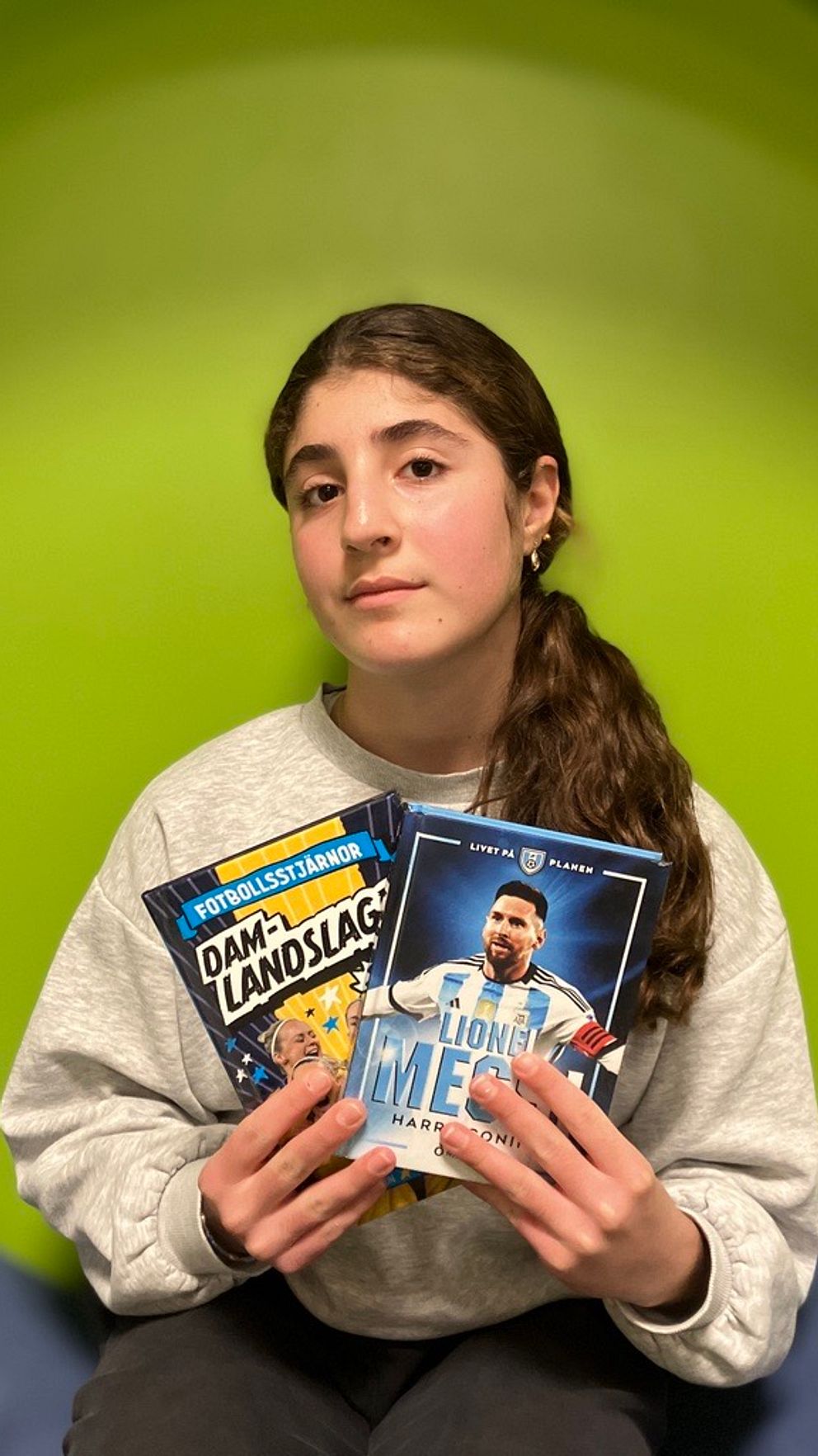 En person håller upp två böcker, en om damlandslaget i fotboll och en biografi om Lionel Messi. Bakgrunden är grön och personen bär en grå tröja.