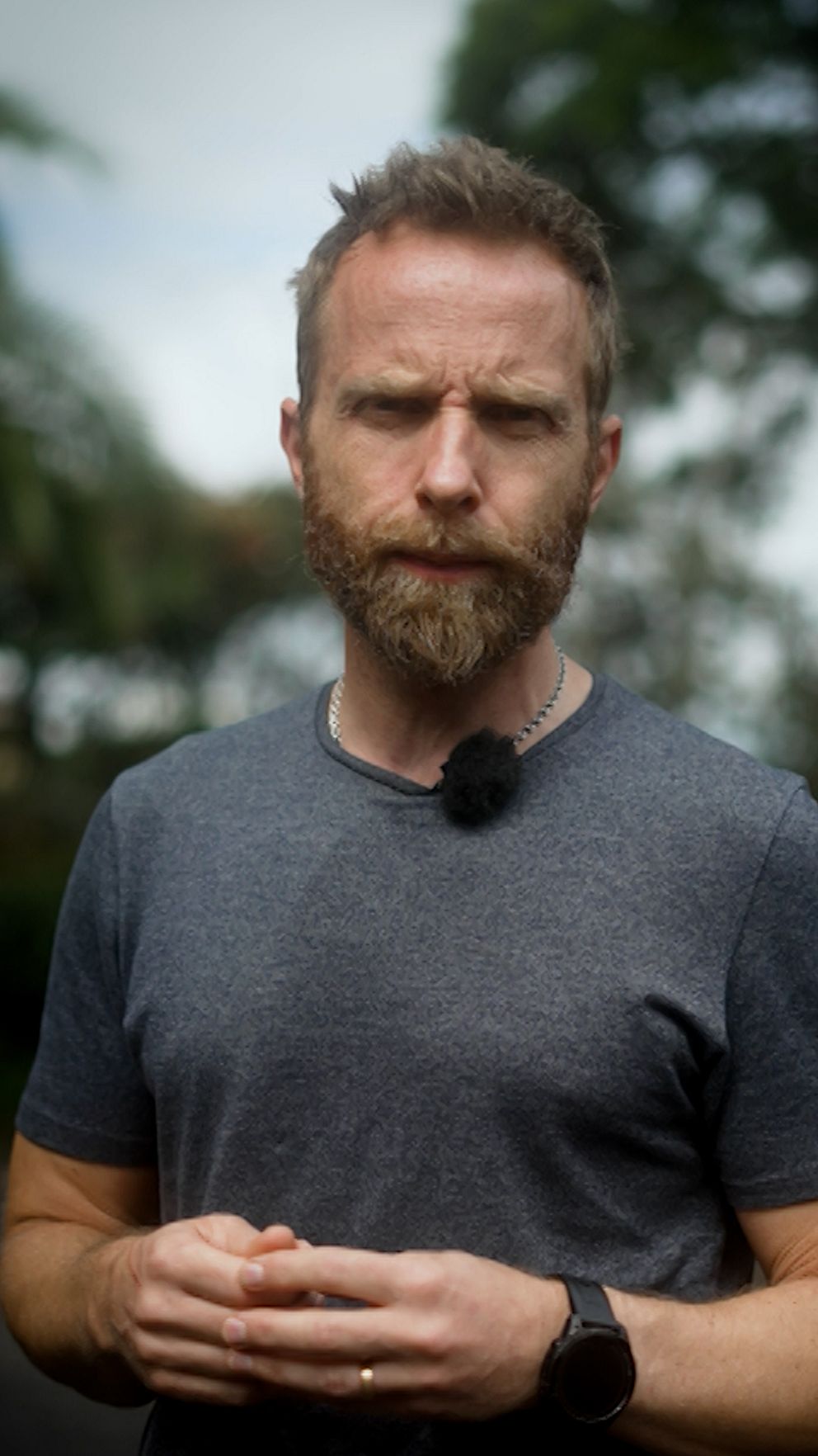 En man i en mörkgrå t-shirt står utomhus framför en suddig lummig bakgrund, bär en mikrofon på kragen och en klocka på vänster handled.