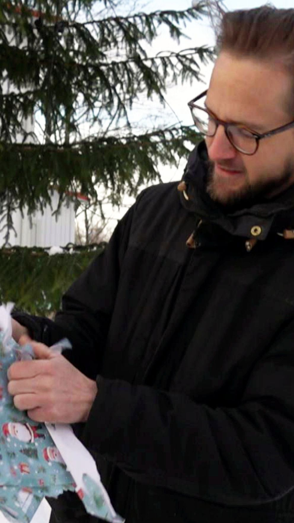 En person i en svart jacka står utomhus i snön och öppnar ett present som är inslaget i papper med snögubbar på. I bakgrunden syns en grön gran och snötäckta landskap.