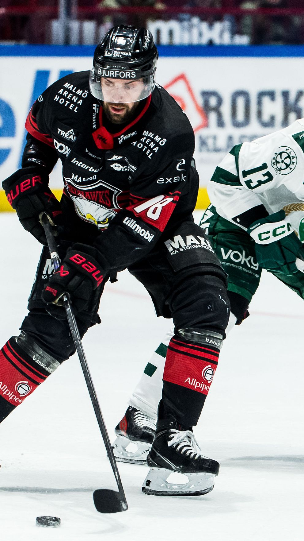 Ishockeyspelare i röd och svart dräkt dribblar pucken förbi motståndare i grön och vit.