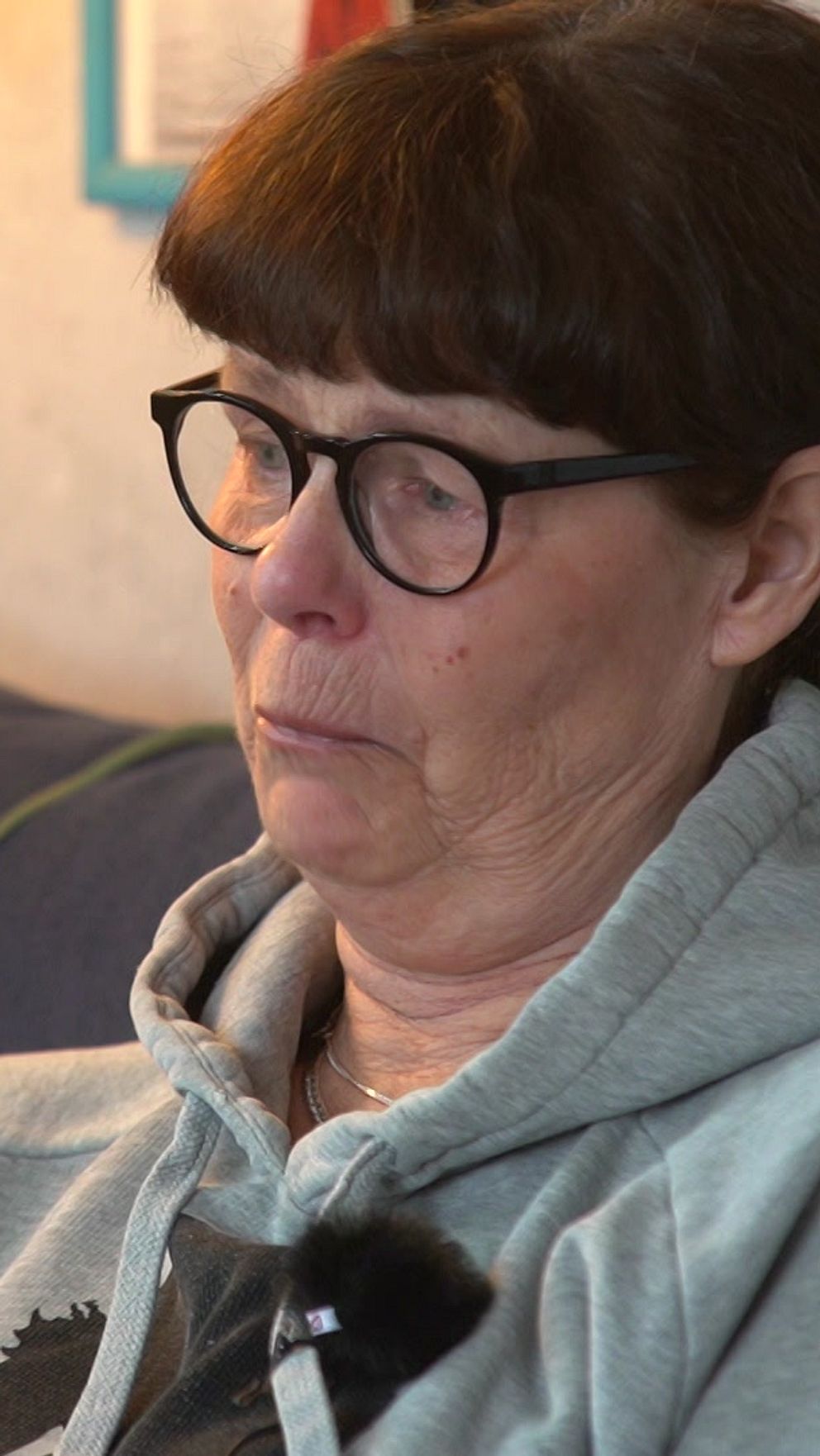 En person sitter på en soffa och bär en grå huvtröja med ett tryck av ett svart träd. I bakgrunden finns en ljus gul vägg med en inramad tavla och en lampa.