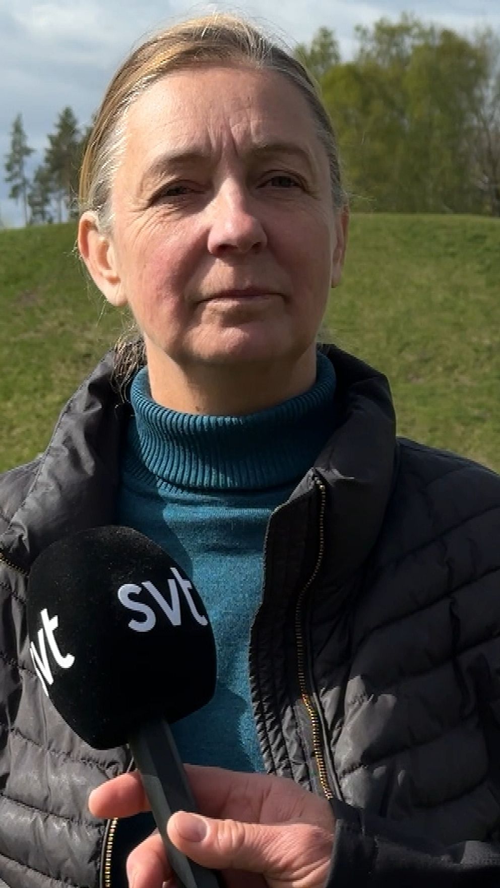 Reporter håller SVT-mikrofon framför person på historisk plats med gravhög och stenar i bakgrunden.