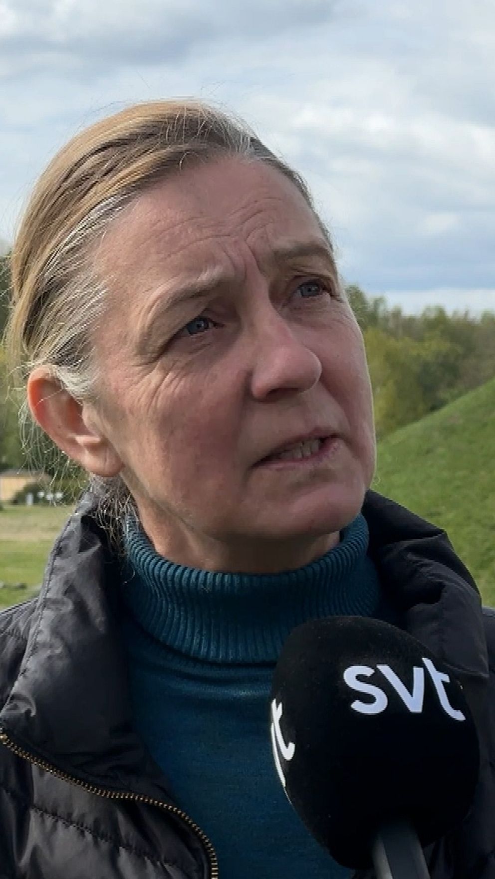 Person intervjuas av SVT på fornlämningsområde med stenar och lantliga hus i bakgrunden.